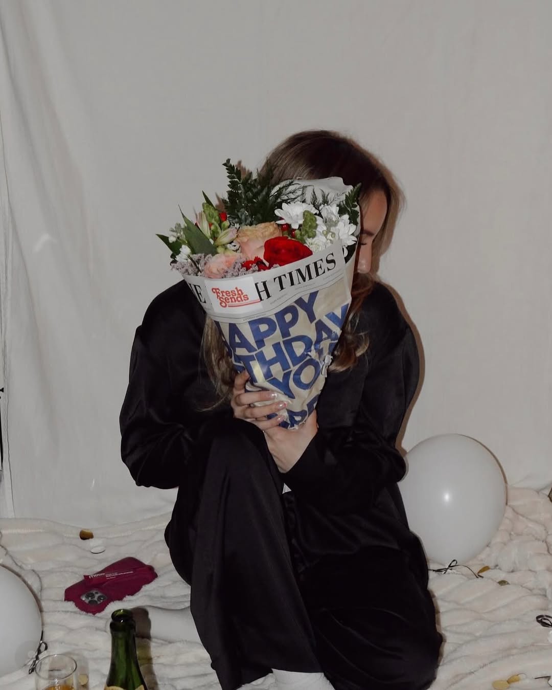 Woman holding a Fresh Sends birthday flower bouquet wrapped in newspaper-style paper, standing indoors with colorful balloons in the background.-318214603119