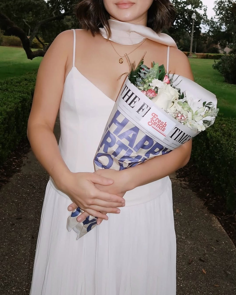 A woman in a white dress holds a bouquet of birthday flowers from Fresh Sends wrapped in a ‘Happy Birthday’ wrapper.