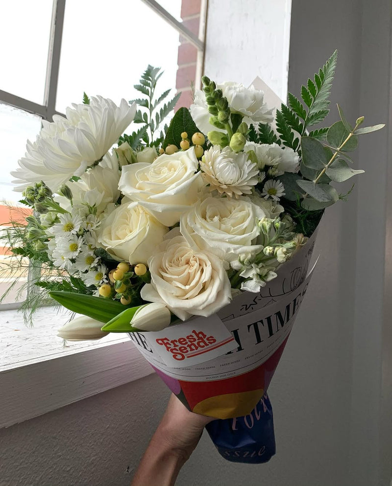 Hand holding a Fresh Sends bouquet of white roses and mixed white flowers in front of a window.