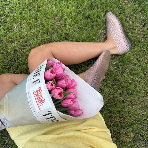 A person in a yellow dress lies on the grass with a bouquet of pink tulips.