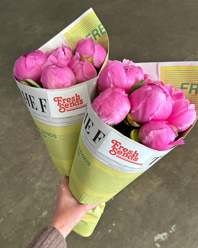 A hand holds two "Fresh Sends" bouquets of vibrant pink peony buds wrapped in yellow paper.-317924933999