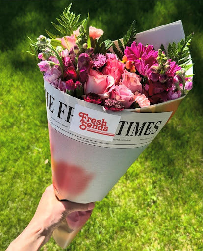 A hand holding a Fresh Sends pink and purple flower bouquet on green grass.-317693722991