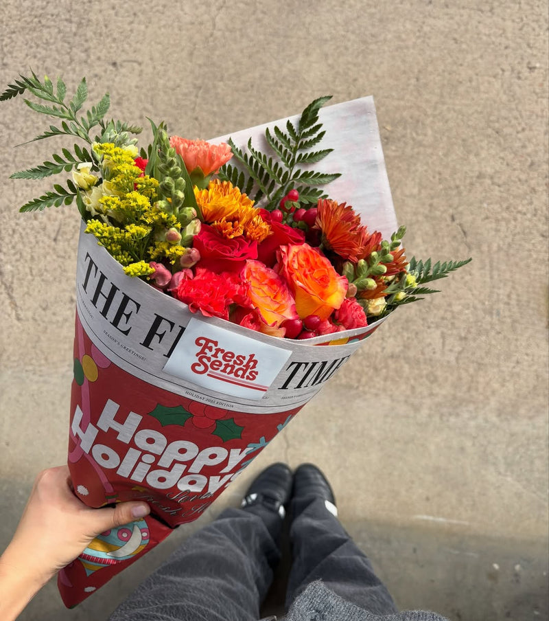 A person holding a Fresh Sends bouquet wrapped in a “Happy Holidays” newspaper-style sleeve.
