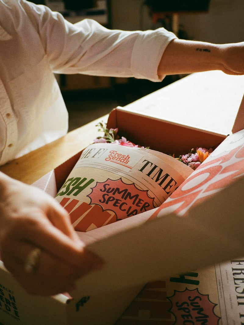 A person opens a Fresh Sends delivery box to reveal a bouquet of flowers wrapped in paper labeled “Fresh Sends Summer Special.”