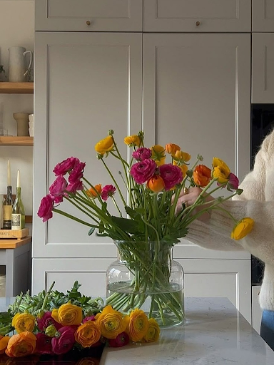 Person arranging yellow, pink, and orange ranunculus stems from Fresh Sends in a clear glass vase on a tabletop.