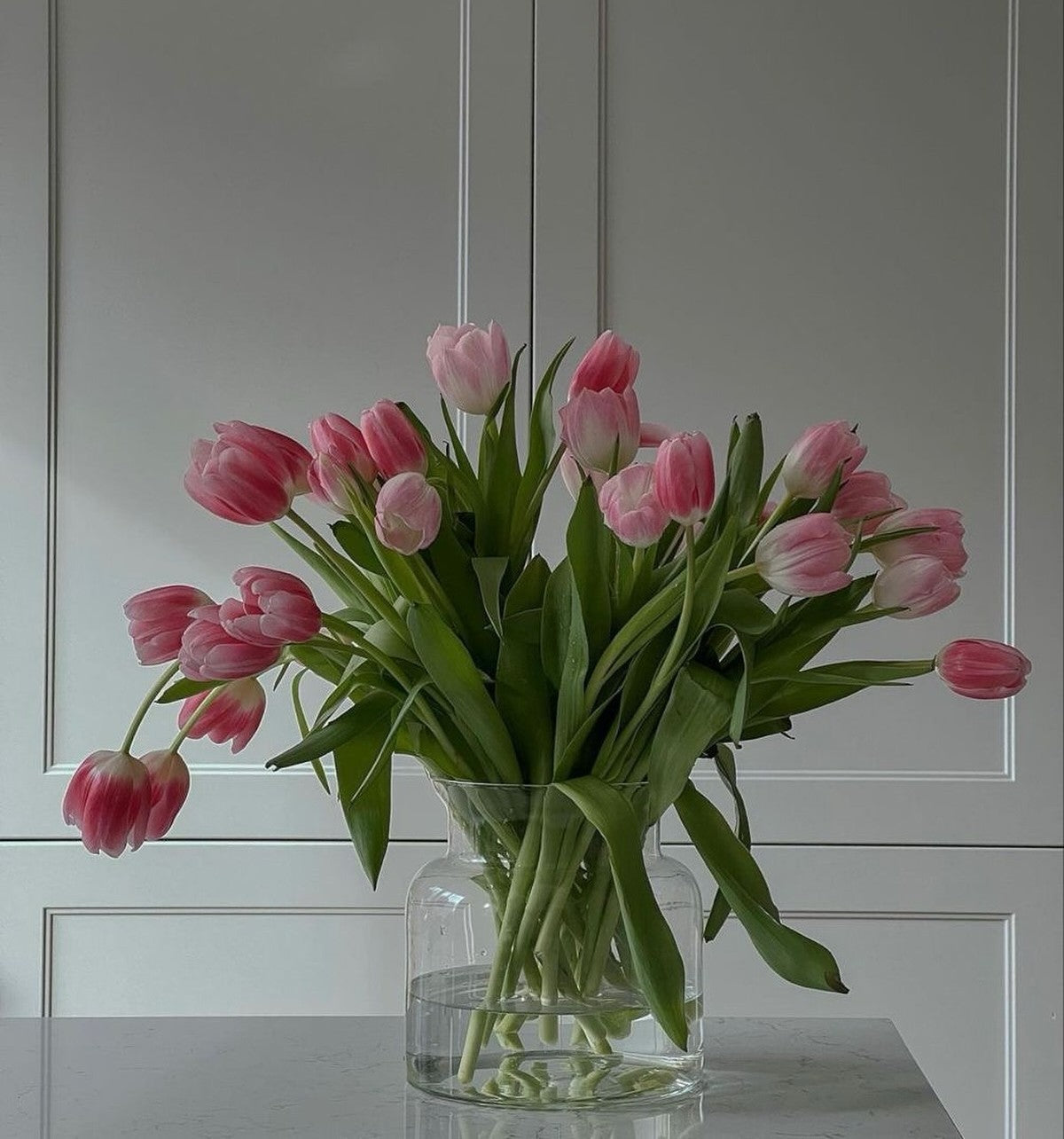 A glass vase filled with water and Fresh sends pink tulips and green leaves on a table.-319060476271