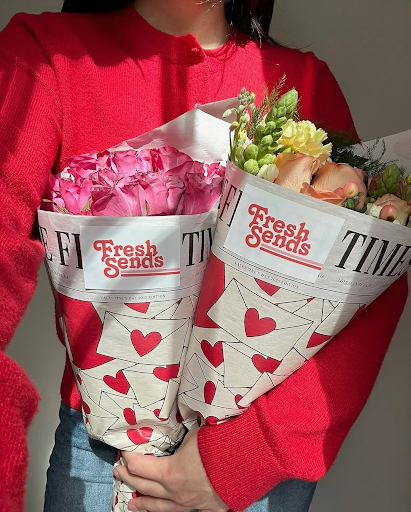 Person in a red sweater holding two bouquets of roses wrapped in ‘Fresh Sends’ paper with hearts.-317656367471