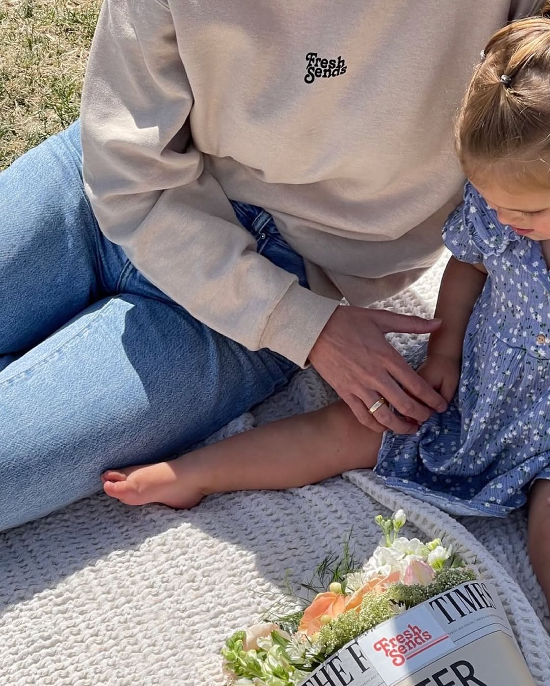 Mother and child sitting on a picnic blanket with a wrapped flower bouquet, sharing a relaxed outdoor moment.-321113063791
